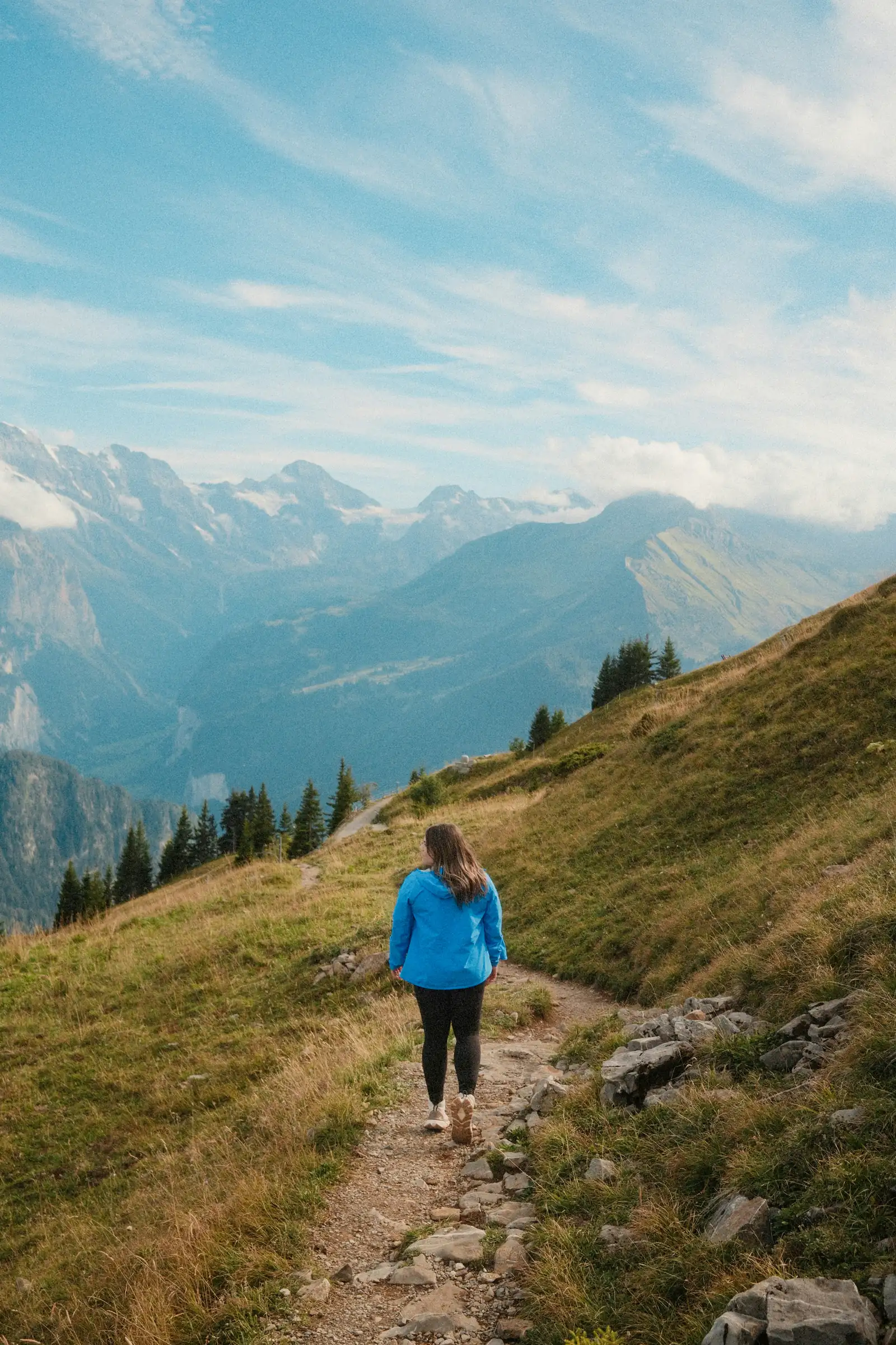 A woman hiking a mountain trail, looking out toward the peaks ahead